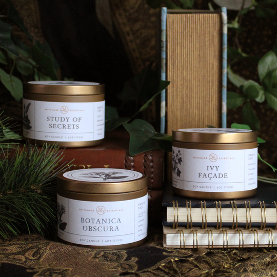 Three candles with labels on a textured surface with books and greenery
